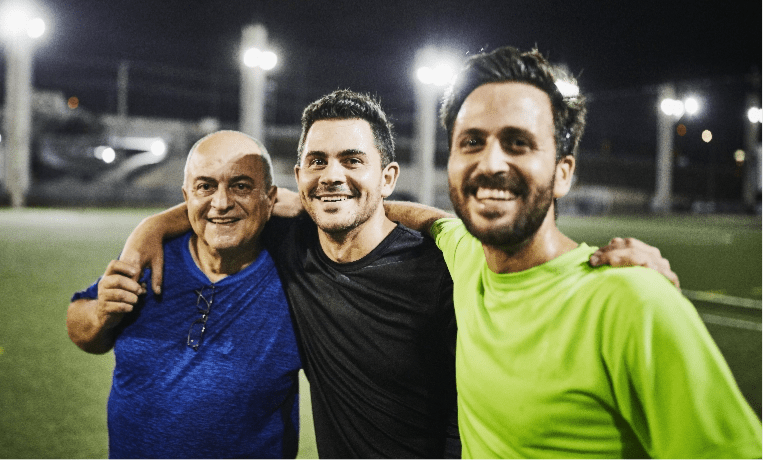 Dos hombres sonríen y abrazan a su padre luego de una partida de fútbol.