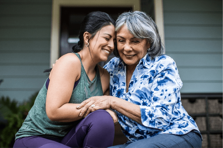 Madre e hija sonríen mientras se abrazan sentadas frente a una casa.